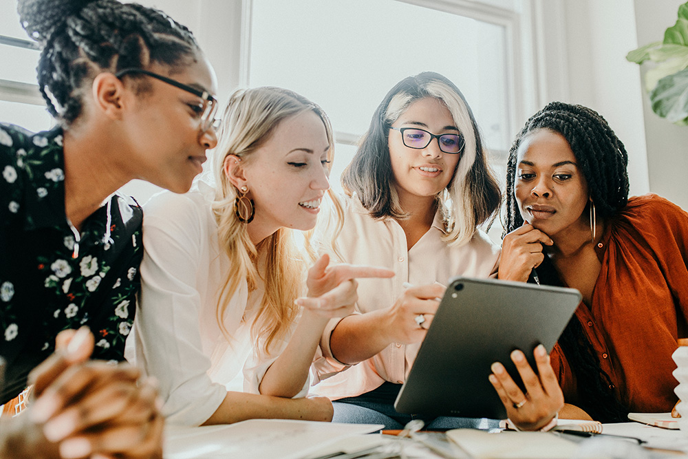 four women in business clothes review a tablet device during a meeting